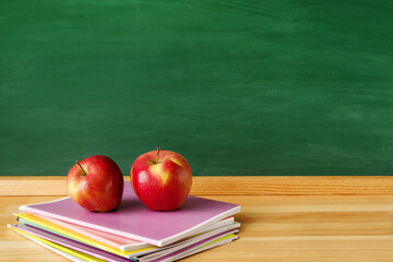 Back to school background. Two red apples and a stack of bright notebooks on a wooden table against the background of a green school board, close-up, copy space.