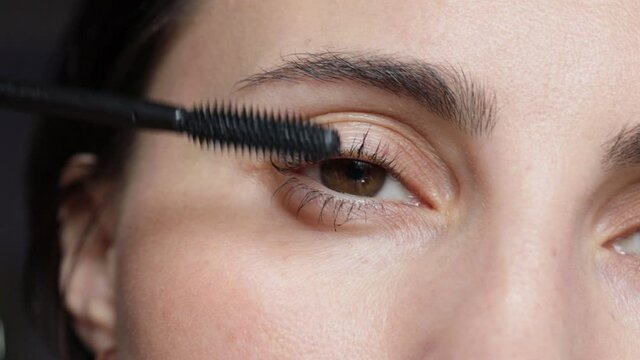 Woman is painting lashes using mascara and brush, closeup face view. Female with brown eyes is doing makeup. Everyday routine for woman.