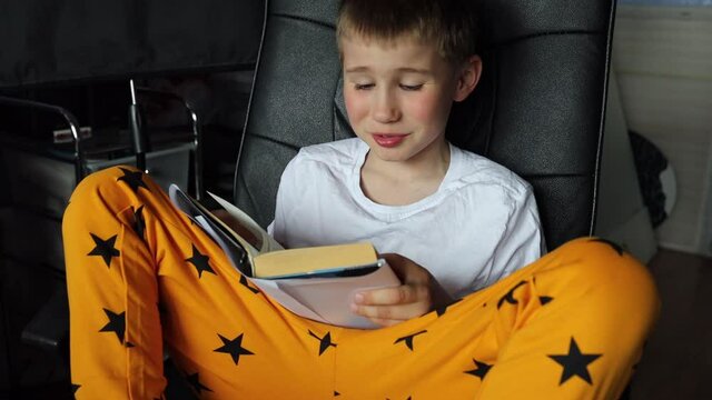 Close Up view of schoolboy sitting on chair at home. Boy reading paper book aloud, turning pages, studying at home