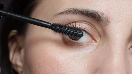 Woman is painting lashes using mascara and brush, closeup face view. Female with brown eyes is doing makeup. Everyday routine for woman.