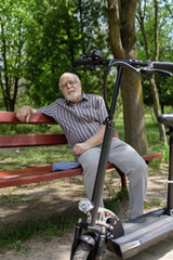 Fototapeta na wymiar A positive, elderly man went for a walk to the park, stopped and sat down to rest on a bench next to an electric scooter. Summer, sunny day. Healthy lifestyle of the elderly.
