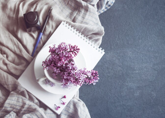 Spring vintage background. Lilac flowers in a white cup, with a white notebook, black ink and a pen for drawing on a background of delicate draped fabric on a gray table, top view, copy space, toned.