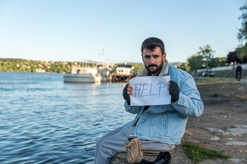 Young depressed sick alcoholic homeless man sitting in the dockyard drinking alcohol drink and holding help paper sign hoping than someone will help with some pocket money, social documentary concept