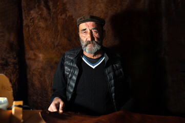 Fototapeta na wymiar Portrait of senior craftsman sitting at the desk in his workshop working with cow skin looking at camera with serious face. Leather background.