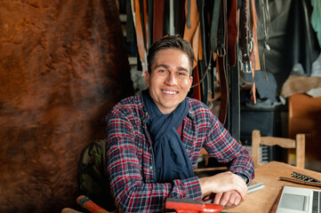 Portrait of confident smiling young male sitting at the desk in his laboratory looking at the camera. Work with genuine leather, handicraft and home hobbies concept