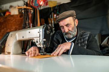 Old hipster bearded man with flat cap working with leather using a sewing machine. Craftsmanship concept.