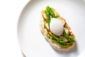 Tasty morning bruschetta with cream cheese, mashed avocado, roasted chicken slices, asparagus and poached egg on white plate isolated. Nutritious brunch recipe. Homemade food on withe background.