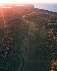 Winding river flowing through forest towards sea. Aerial sunset. Sunlight rays. Baltic sea, Latvia.