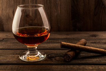 Glass of brandy and two different cuban cigars on a old wooden table. Focus on the cuban cigars