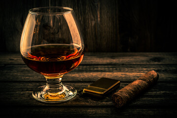 Glass of brandy and cuban cigar with golden lighter on an old wooden table. Focus on the cuban cigar