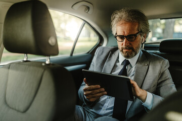 Middle aged businessman using tablet and earphones, having video call while driving in car.