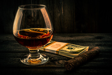 Glass of brandy with pack of dollars and cuban cigar on an old wooden table. Focus on the glass of...
