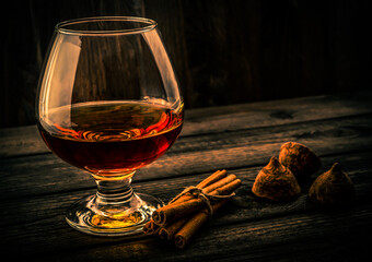 Glass of brandy and a couple of chocolate truffles with cinnamon sticks tied with jute rope on an old wooden table. Focus on the glass of brandy