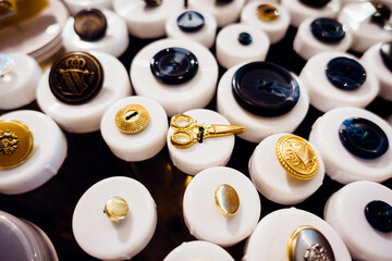 Many tailor accessories are displayed on the table, with black buttons, golden scissors, royal emblems, round shiny surfaces, etc.