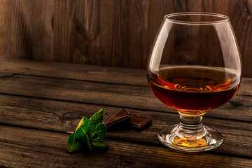 Glass of brandy and a chocolate with mint sprig on an old wooden table. Angle view