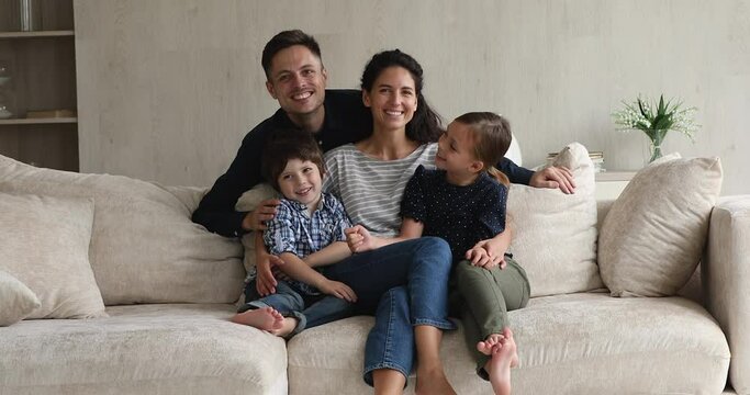 Beautiful happy spouses with preschool children sit on sofa pose on camera at home. Happy homeowners young married couple and kids portrait. Medical insurance cover for full family, bank loan concept