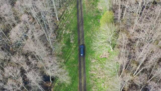 Aerial above shot following a car driving through dirt road surrounded by wet forest. Leafless trees around