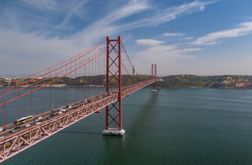 Long red multideck suspension bridge connecting Lisbon to Almada over wide river from drone. Heavy traffic on multilane road.