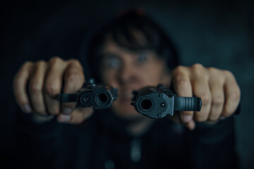 Guy threatens with firearm. Criminal with weapon. Murderer in hood on dark background. Two pistols in man's hands are pointed at camera. Close-up of two gun muzzles.