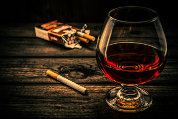 Glass of brandy and cigarette with pack of cigarettes on a old wooden table. Angle view