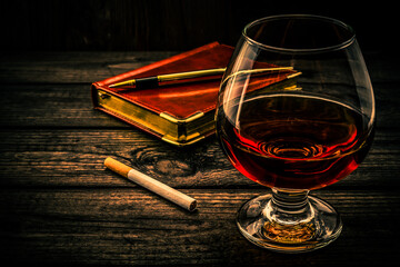 Glass of brandy and cigarette with leather notebook and golden pen on an old wooden table. Angle view