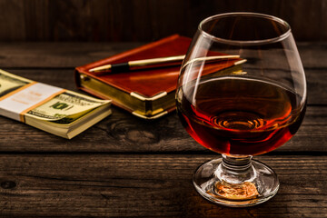 Glass of brandy and leather notebook with golden pen and pack of dollars on an old wooden table. Angle view, focus on the glass of brandy
