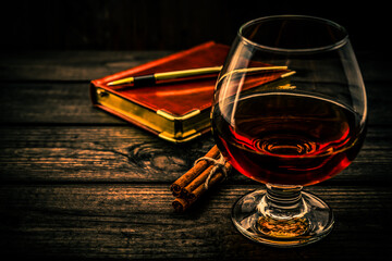 Glass of brandy and leather notebook with golden pen and cinnamon sticks tied with jute rope on an old wooden table. Angle view