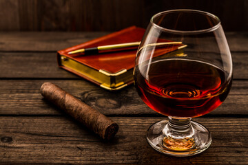 Glass of brandy and cuban cigar with leather notebook and golden pen on an old wooden table. Angle view