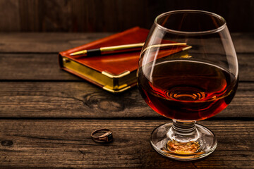 Glass of brandy and leather notebook with golden pen and ring on an old wooden table. Angle view