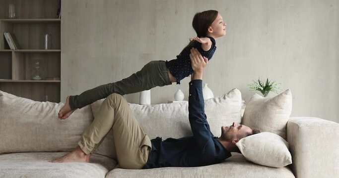 In living room strong dad lying on sofa holding lifting little daughter on outstretched hands, child spread arms like plane wings imagines like fly in air. Sportive family, yoga practice, fun concept