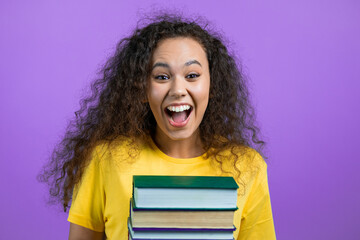 Student girl holds stack of university books from library on violet background in studio. Woman smiles, she is happy to graduate.