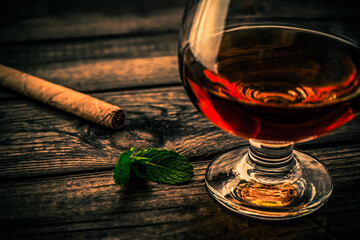 Glass of brandy with mint sprig and cuban cigar on an old wooden table. Close up view, shallow depth of field