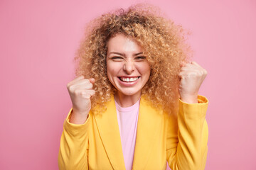 Cheerful overjoyed curly haired businesswoman clenches fists like champion celebrates victory hears...