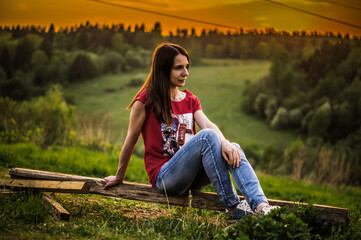 Woman is sitting on hills top against colorful sunset. Enjoying and relaxing in nature. Girl sitting on a wooden board alone on sunset background
