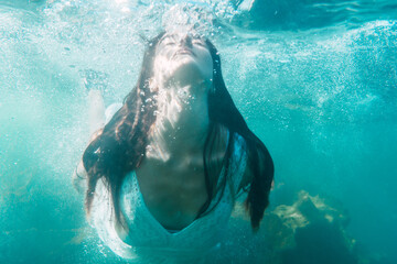 A young Caucasian brunette in a white dress inside the sea, underwater image, summer lifestyle
