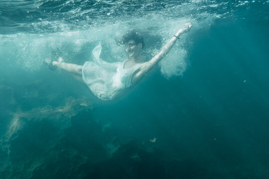 A young Caucasian brunette in a white dress jumping into the sea, underwater image, summer lifestyle