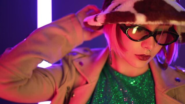 Charismatic woman with pink hairstyle posing in panama hat near led-colorful neon lamps. Hipster trendy girl in sunglasses. Modern model pop outfit, influencer lifestyle. Slow motion.