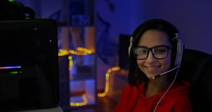 Portrait, happy girl streamer in glasses sits in headphones with a microphone and smiles. Beautiful girl streamer in a computer chair in a dark room. 4k, ProRes