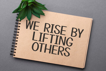 We Rise by Lifting Others text on craft colored notepad and green plant on the dark background