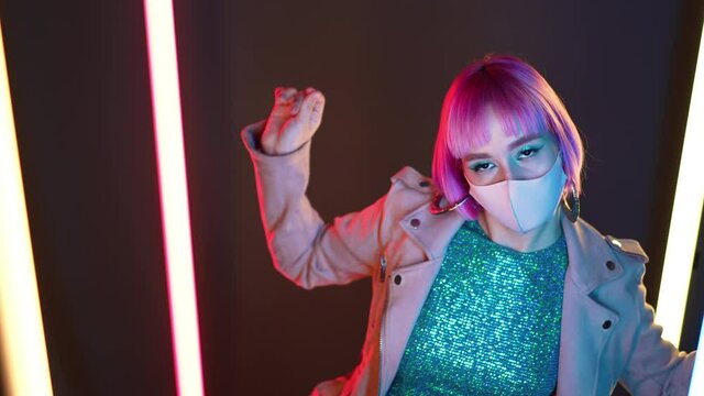 Amazing woman with pink hair and facial protective mask dancing alone between led-colorful bright lamps. Charming lady, night life concept. Modern pop outfit, influencer lifestyle.