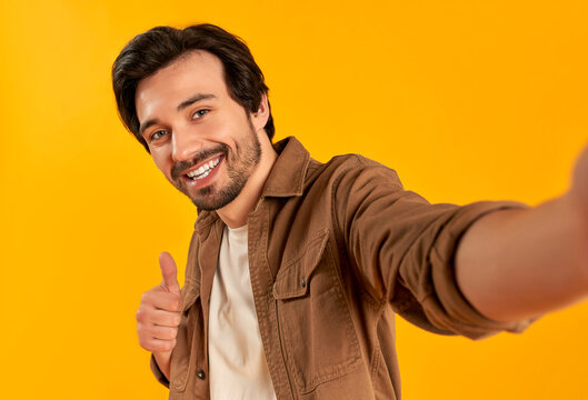 Young bearded man showing thumbs up gesture and making selfie photo on smartphone smiling isolated on orange background.