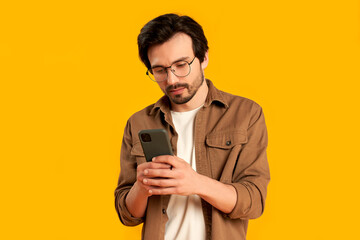 Young bearded man with glasses uses a smartphone, types on the keyboard, communicates isolated on an orange background.