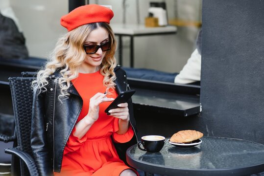 Young stylish woman in red beret having a french breakfast with coffee and croissant sitting oudoors at the cafe terrace