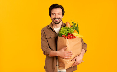 Young bearded man with paper package of vegetables groceries isolated on orange background.