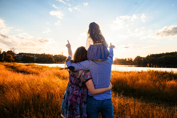 happy family with a little daughter in a field in nature, looking forward, view from the back, in the rays of the sunset - 434616291