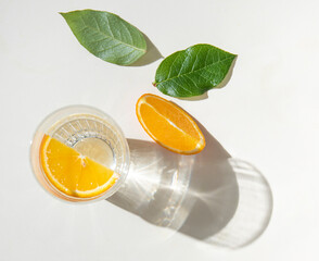 A glass of mineral water and a slice of orange on a light  table with fresh juicy fruit oranges and green leaves with hard shadows. Summer time. Top view