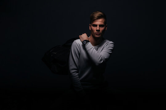 Informally ( casual ) dressed blonde young man with sharp jawline in his 20's posing in a studio in front of a black background while wearing a white sweater and holding a black man bag.