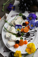 Camembert on a white plate with grape slices, physalis and viola flowers. A light snack in a light dish on a dark table.