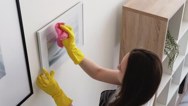 Cleaning woman. Home hygiene. Room service. Work responsibility. Back view woman in apron and yellow gloves clearing dust from picture frame on wall.