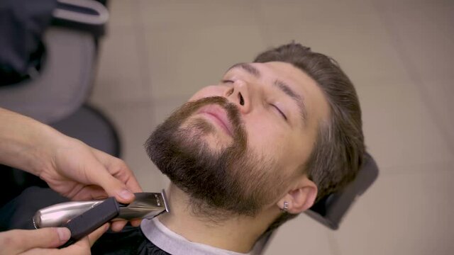 Trimming beard in barbershop. Barber is shaving beard and mustache for handsome customer.